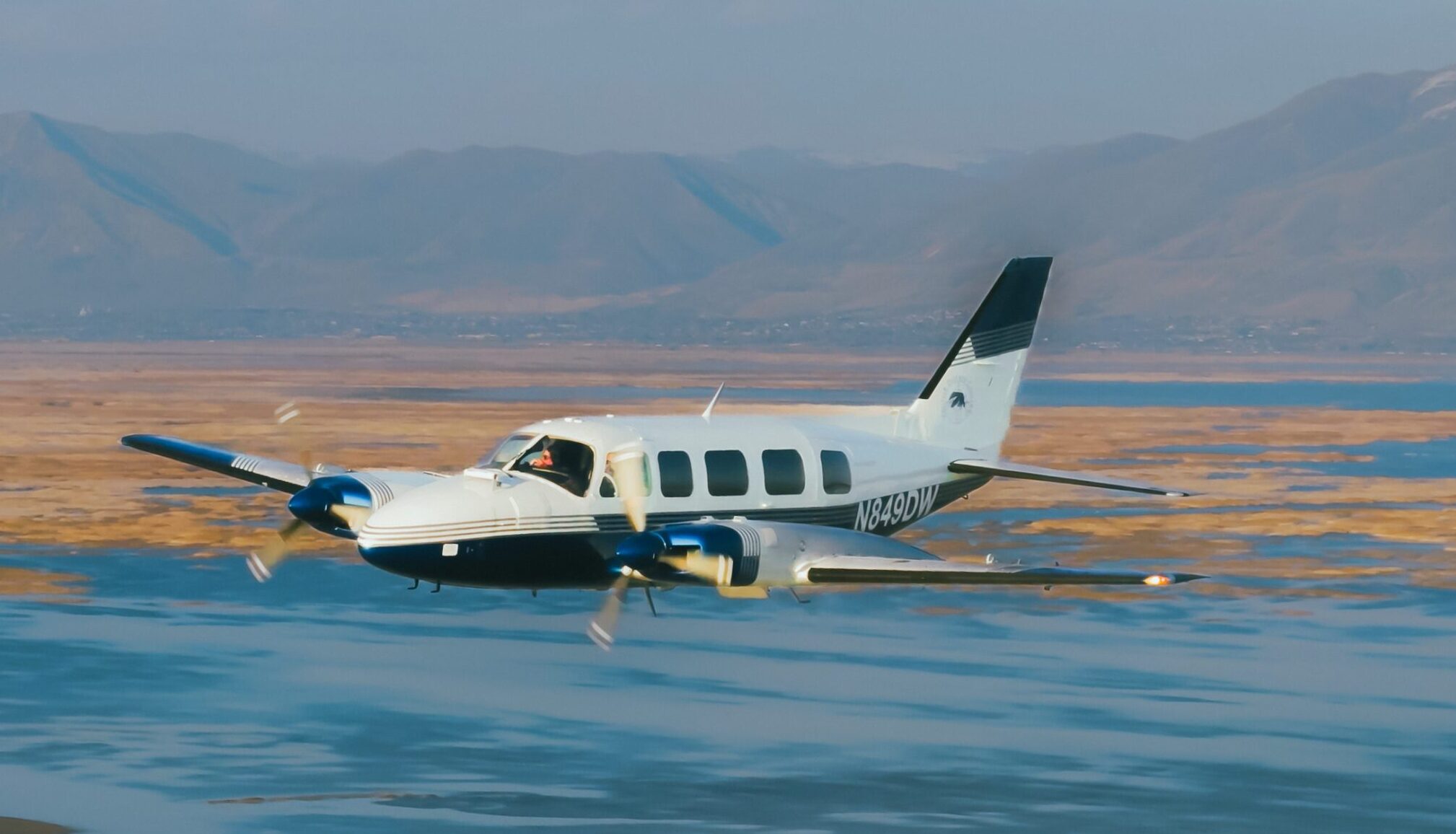White and teal twin-engine aircraft flying over desert landscape with mountains in background