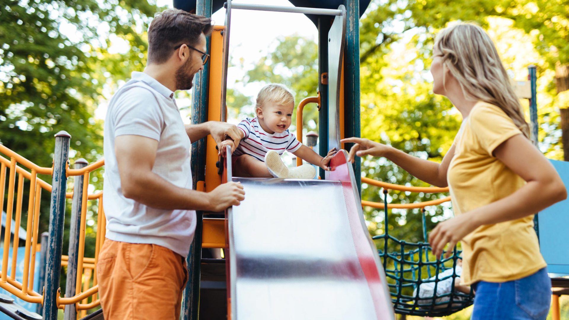 Cheerful parents having fun with their son in the playground. Little boy is sliding family playing on playground structure